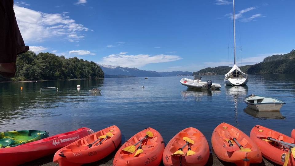 Fila de kayaks naranjas en la orilla de la playa, con botes amarrados en la bahía