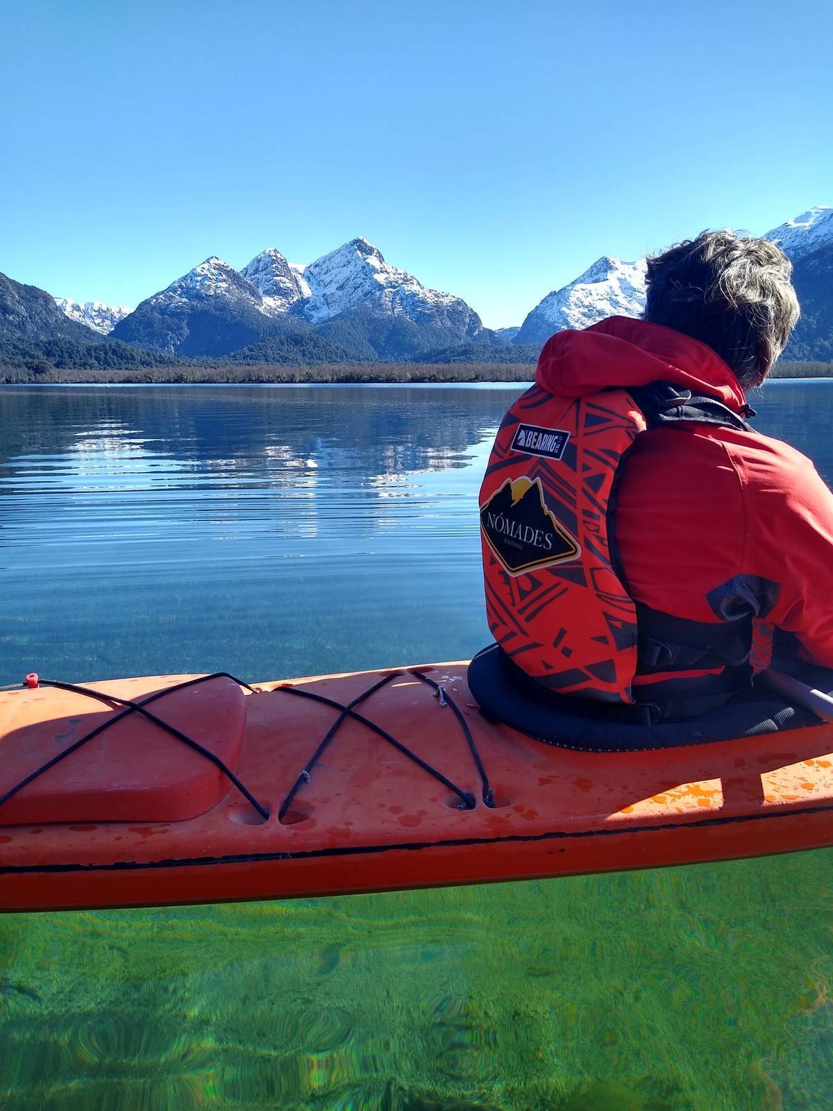 Persona en un kayak rojo con un chaleco salvavidas con el logo de Nómades, frente a montañas nevadas que se reflejan en el lago