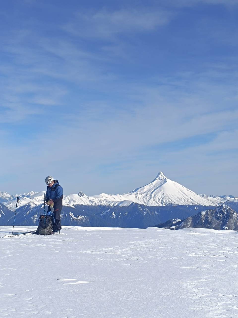 Persona con casco y mochila en una planicie nevada, con un volcán nevado en punta y la cordillera de fondo