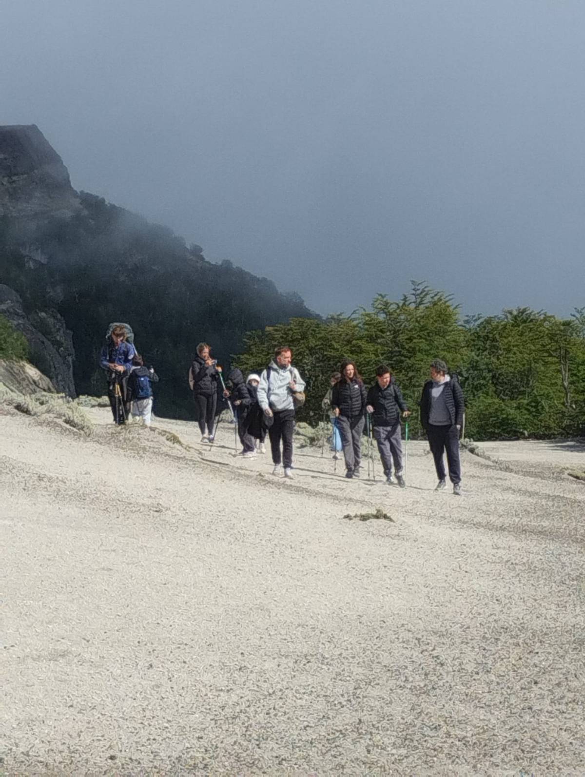 Grupo caminando con bastones de trekking por una ladera de ripio claro, con bosque y niebla detrás