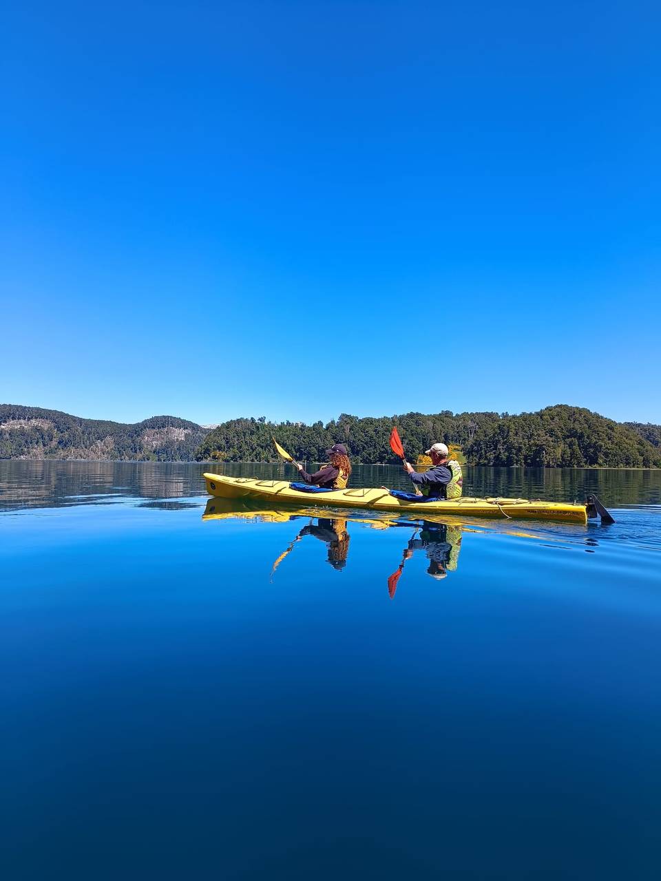Kayak doble amarillo reflejado en el agua calma, frente a una isla cubierta de bosque