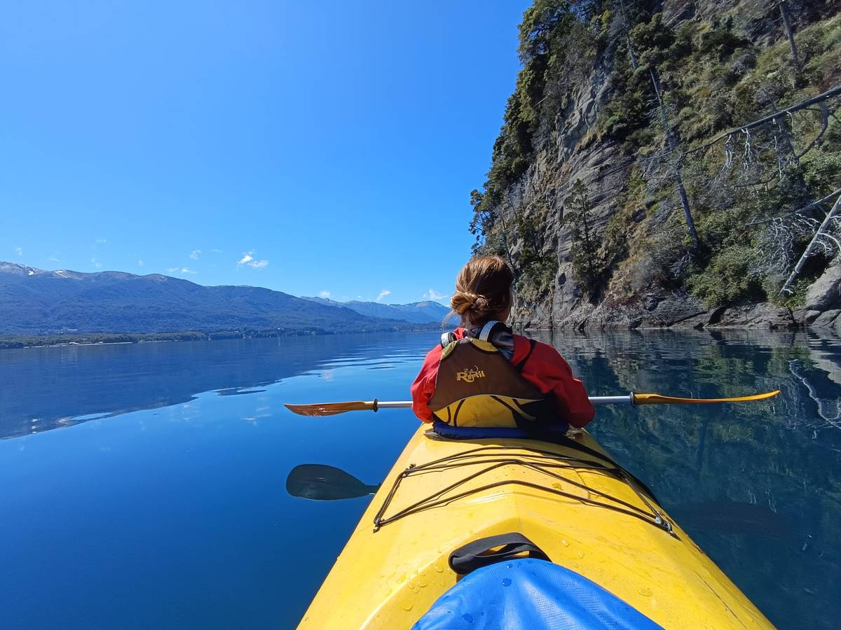 Vista desde un kayak amarillo: una persona con campera roja rema junto a un paredón de roca con árboles, sobre un lago azul y calmo