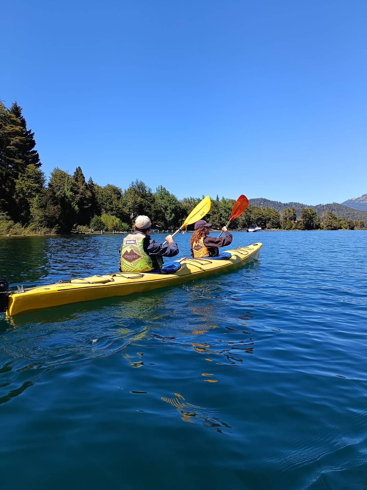 Dos personas remando en un kayak doble amarillo cerca de una costa con bosque, bajo el cielo azul