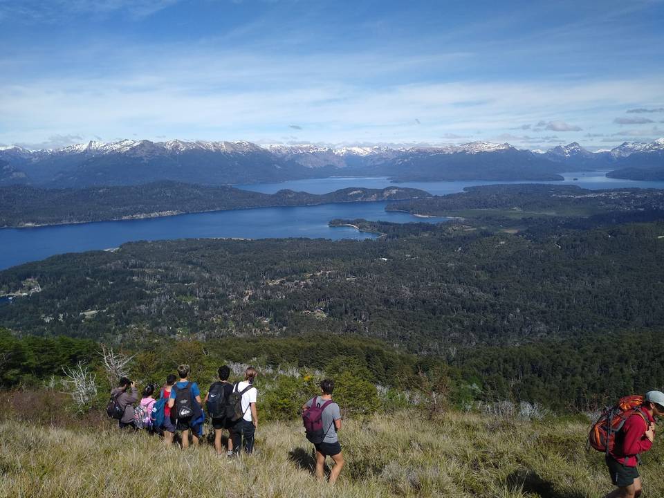 Grupo de caminantes con mochilas en una ladera con pasto, mirando el lago Nahuel Huapi, el bosque, Villa La Angostura y la cordillera nevada